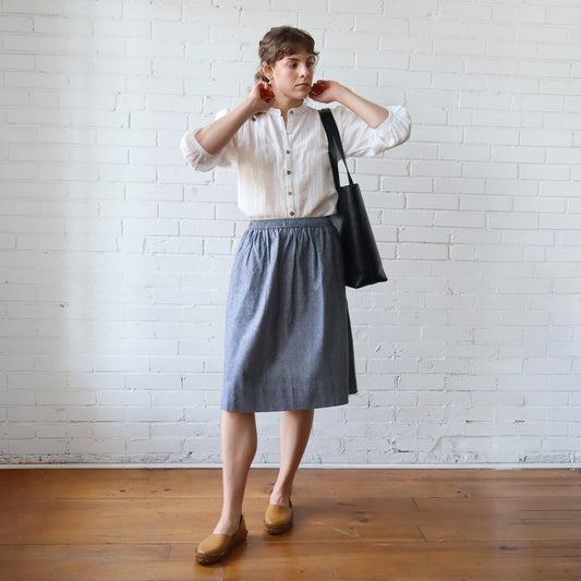 Person wearing a white shirt and gray skirt with a black bag against a white brick wall.