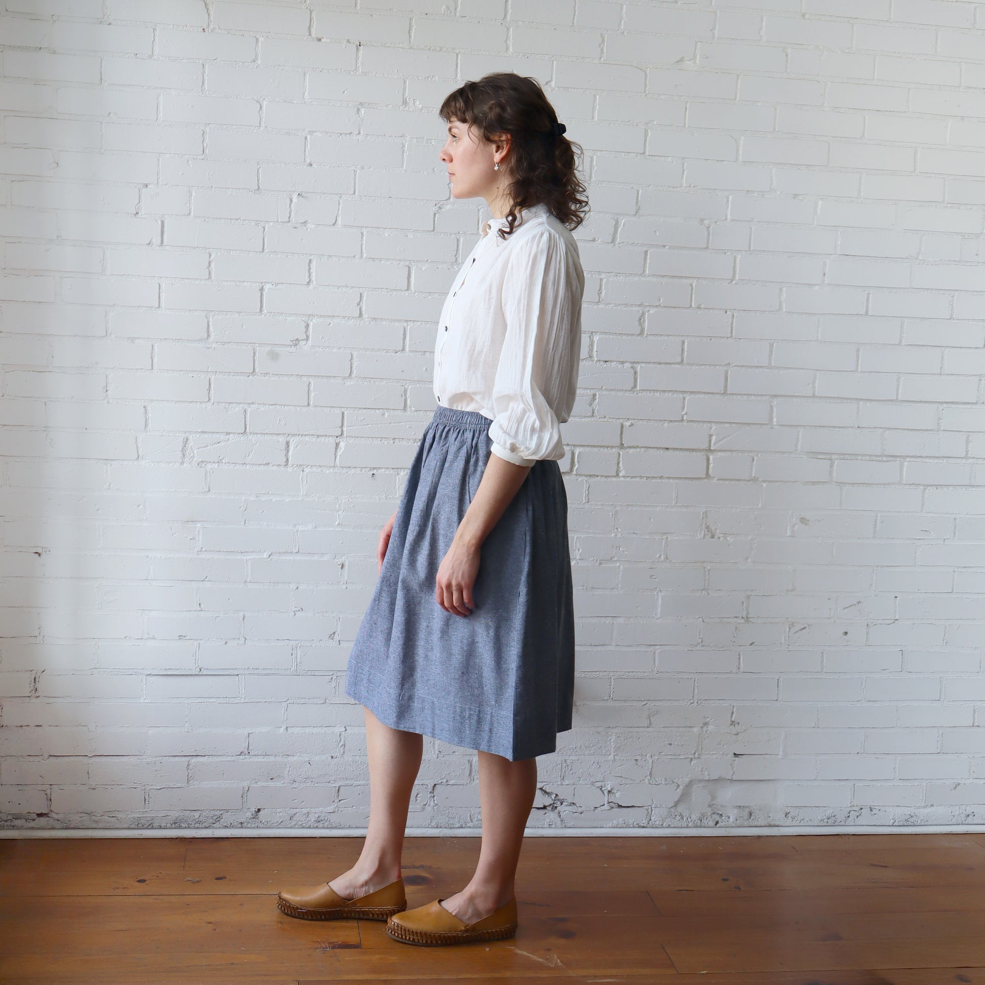 Woman wearing a white blouse and blue skirt standing against a white brick wall.
