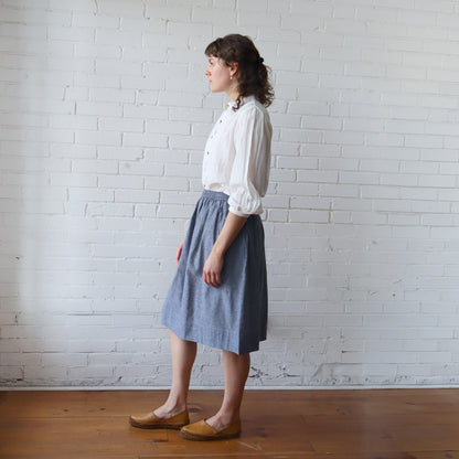 Woman wearing a white blouse and blue skirt standing against a white brick wall.