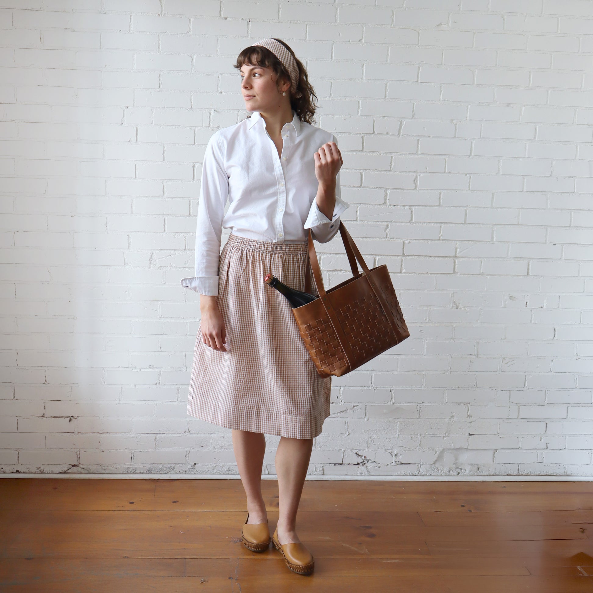 Woman holding a brown tote bag against a white brick wall.