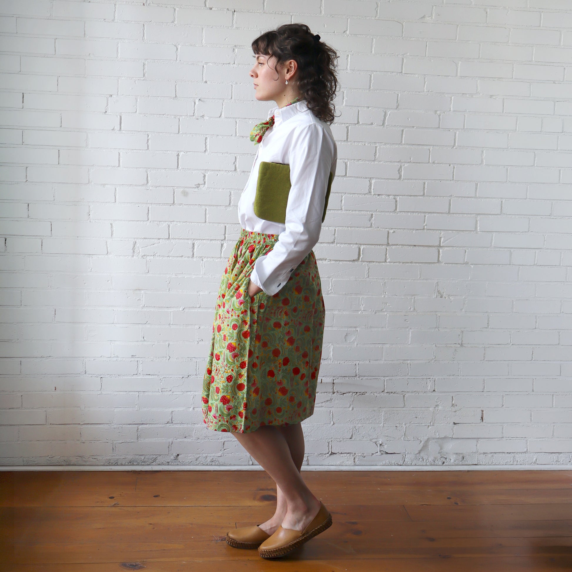 Person wearing a white shirt and patterned skirt against a white brick wall.