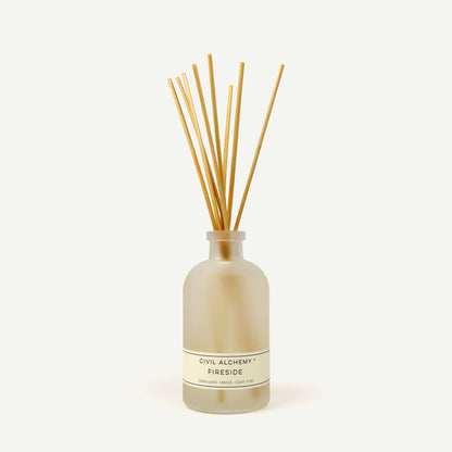 Reed diffuser with wooden sticks in a clear bottle labeled 'Civil Alchemy Fireside' on a white background