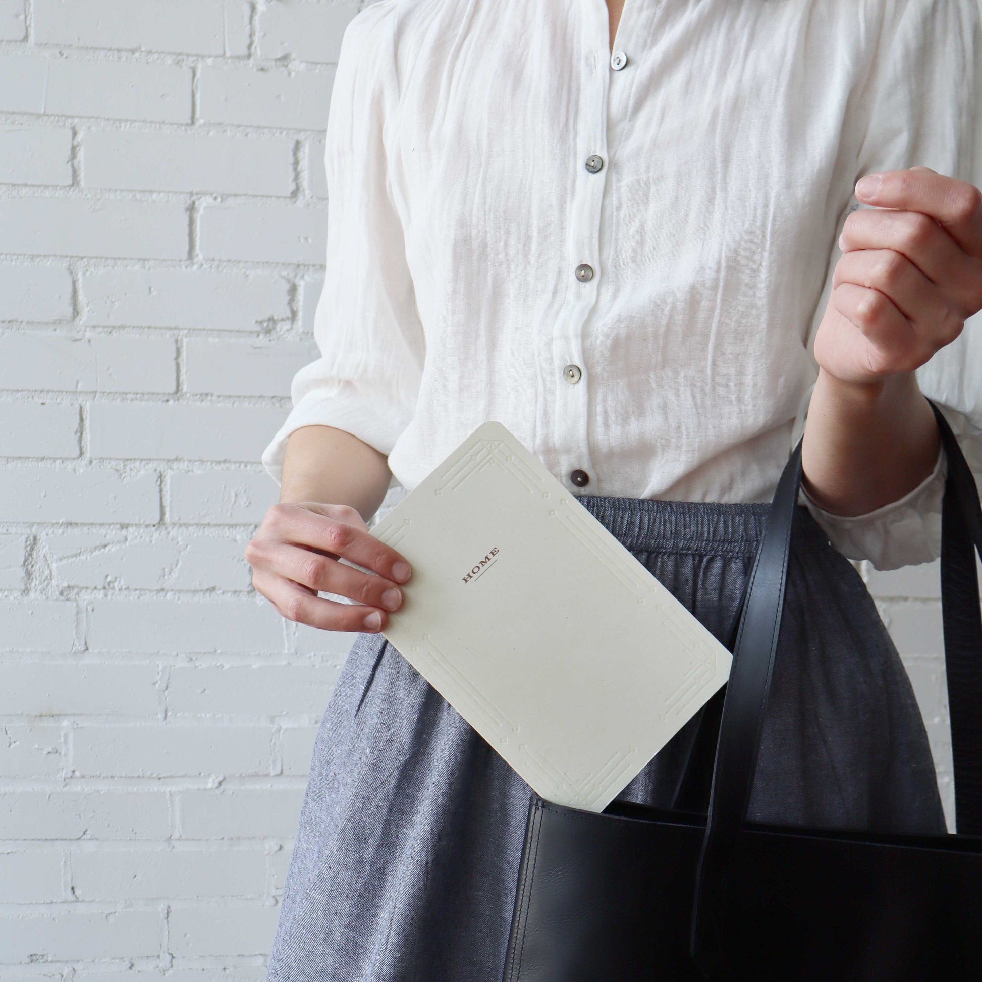 A person wearing a white button-up shirt and a gray skirt holds a cream-colored notebook with the word 'HOME' visible on the cover, while carrying a black tote bag against a white brick wall background.