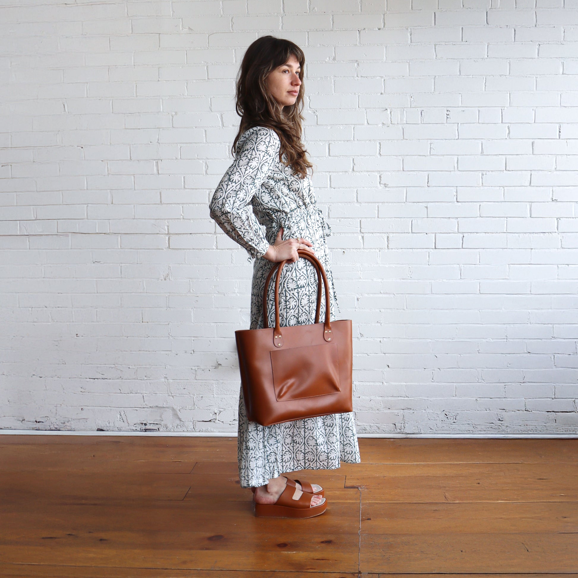 Woman holding a brown leather tote bag against a white brick wall.