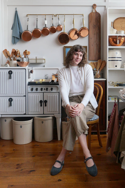 A woman sits on a wooden chair in a rustic kitchen setting, wearing a white cardigan over a cream top, beige trousers, and dark blue Mary Jane shoes. The background features a white vintage-style stove, hanging copper pots, wooden cutting boards, and white shelving units.