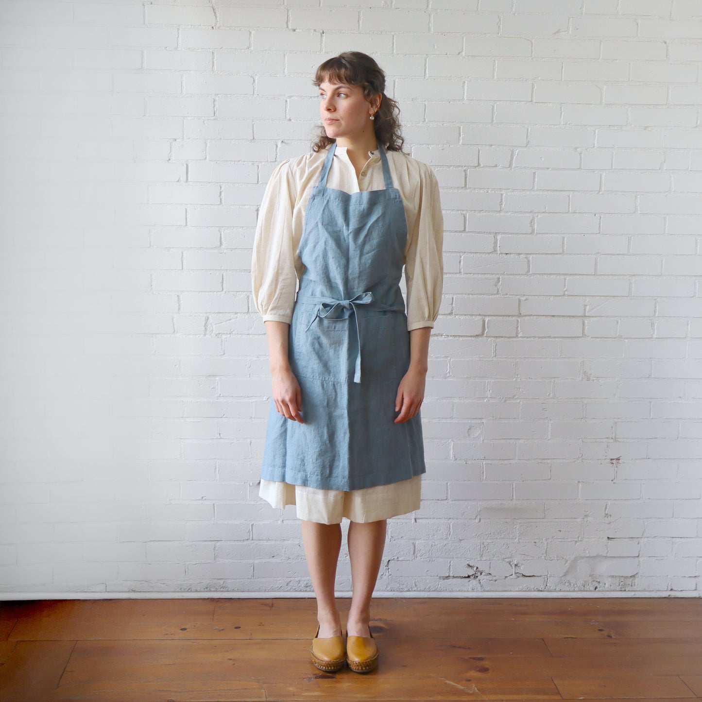Woman wearing a blue apron over a white shirt and skirt against a white brick wall.
