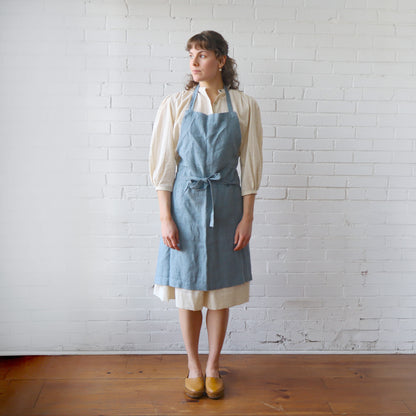 Woman wearing a blue apron over a white shirt and skirt against a white brick wall.