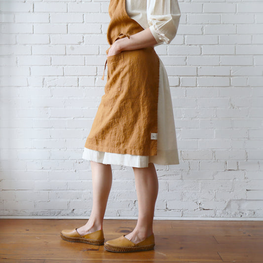 Person wearing a mustard yellow apron over a white dress against a white brick wall.