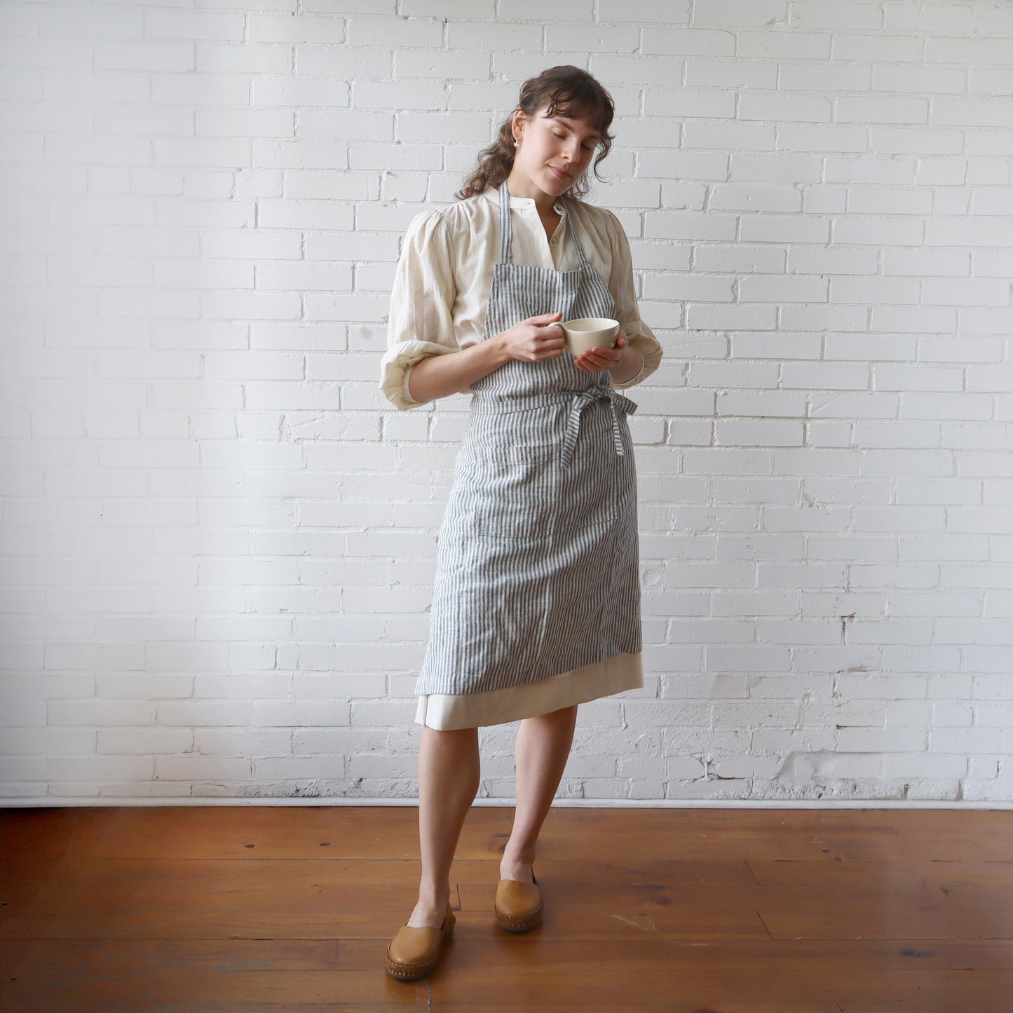 Woman wearing a striped apron holding a cup against a white brick wall.