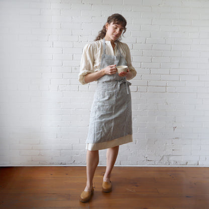 Woman wearing a striped apron holding a cup against a white brick wall.