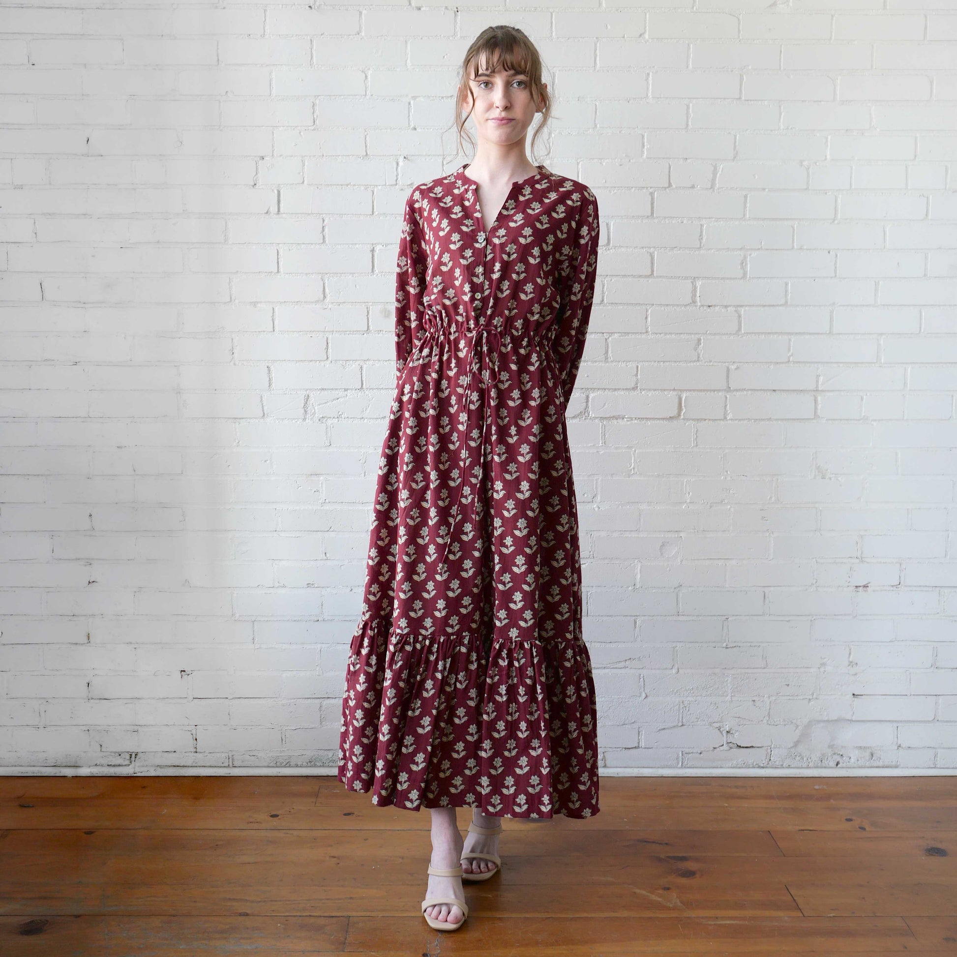 Woman wearing a long burgundy dress with a floral pattern against a white brick wall.