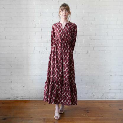 Woman wearing a long burgundy dress with a floral pattern against a white brick wall.