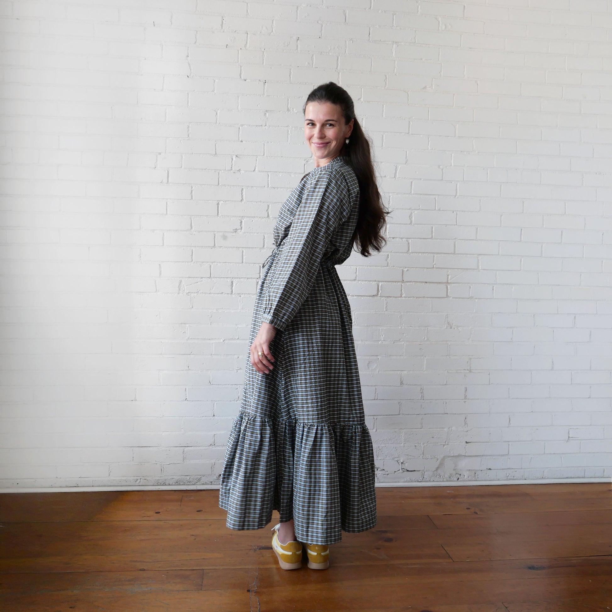 Woman in a checkered dress standing against a white brick wall.