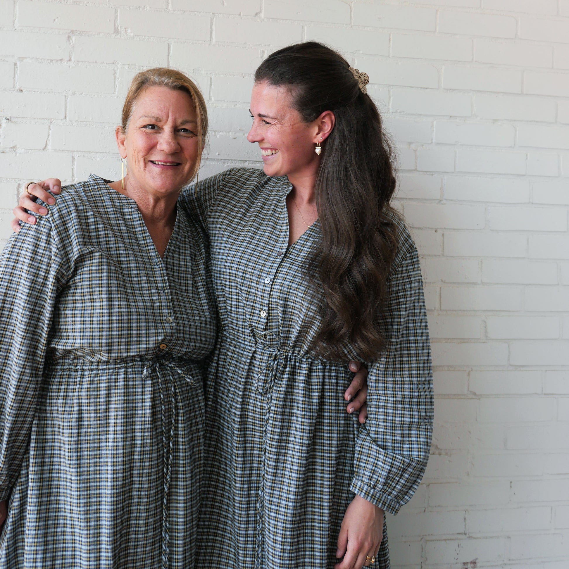Two women wearing matching plaid dresses standing against a white brick wall.