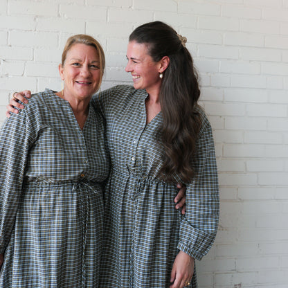 Two women wearing matching plaid dresses standing against a white brick wall.