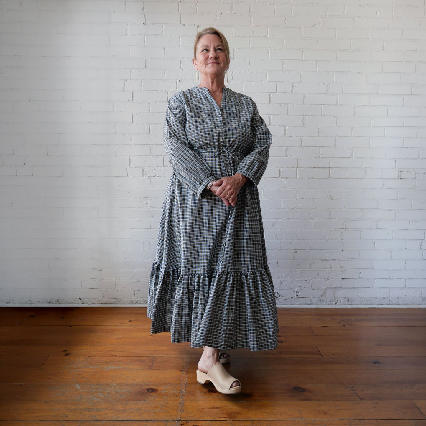 Woman wearing a long, checkered dress standing against a white brick wall.