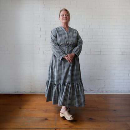 Woman wearing a long, checkered dress standing against a white brick wall.