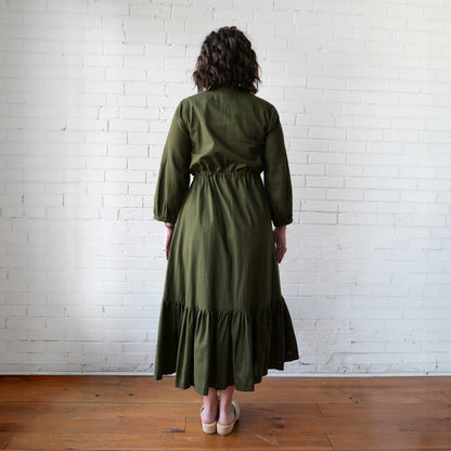 Person wearing an olive green dress standing against a white brick wall.