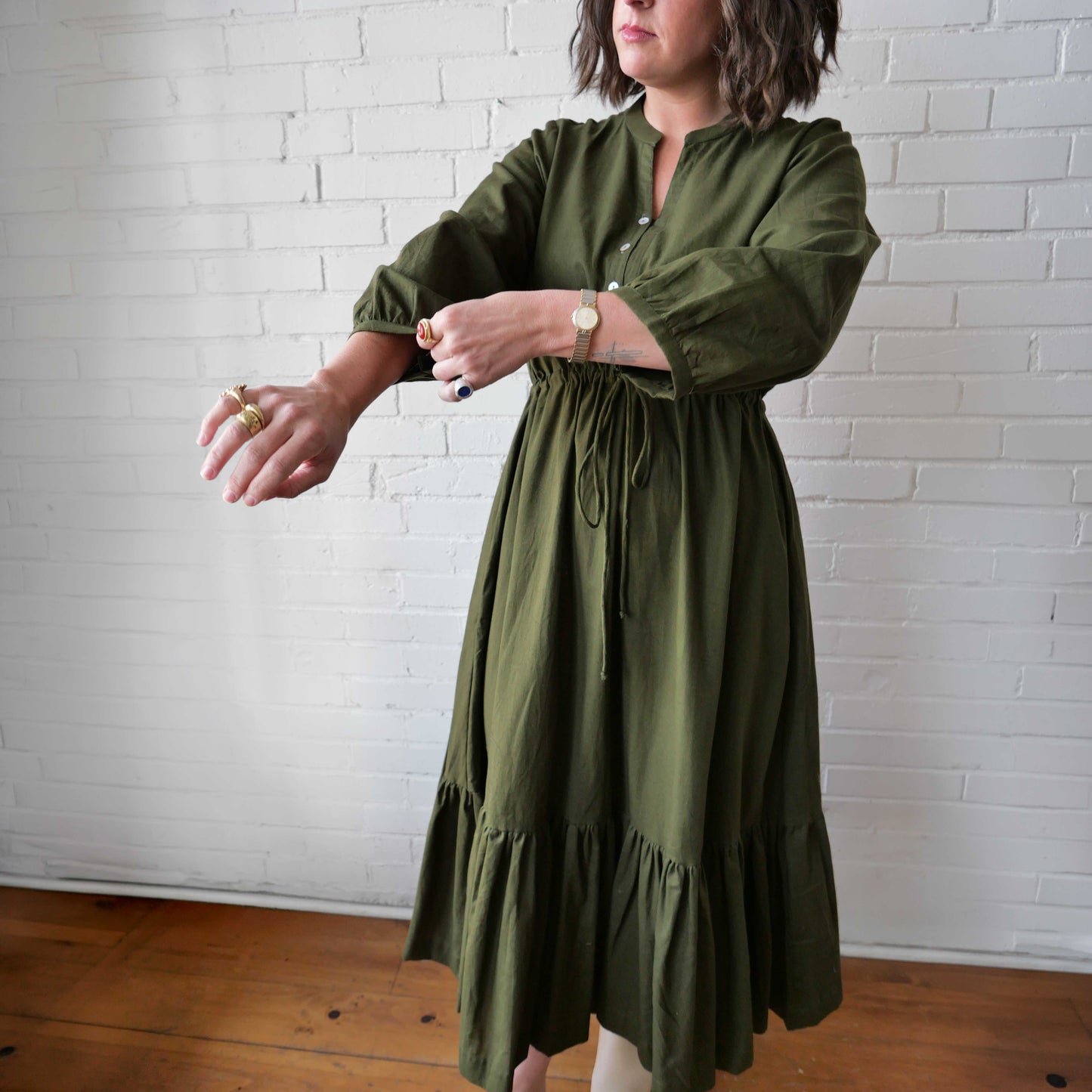 Woman wearing an olive green dress against a white brick wall.