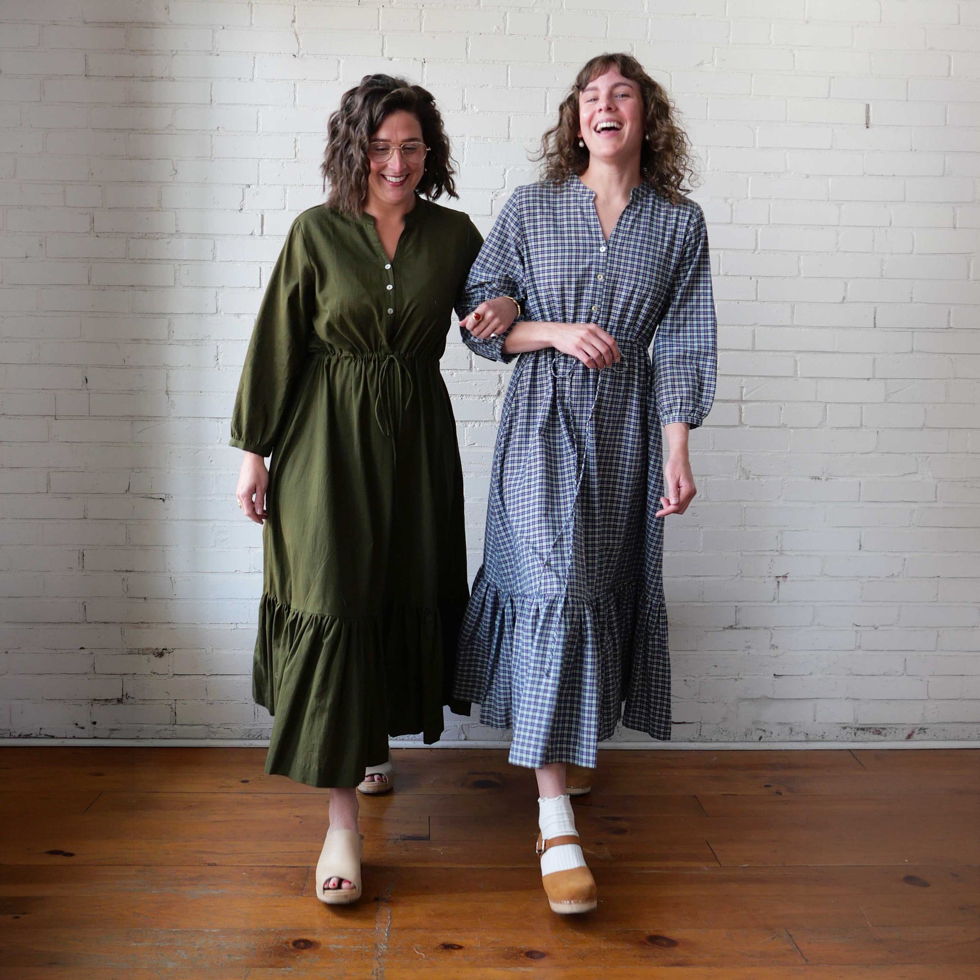 Two women standing side by side wearing long dresses against a white brick wall.