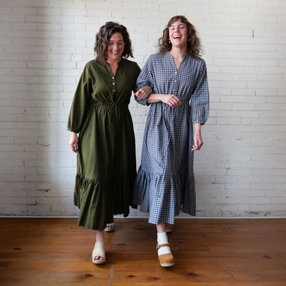Two women standing side by side wearing long dresses against a white brick wall.