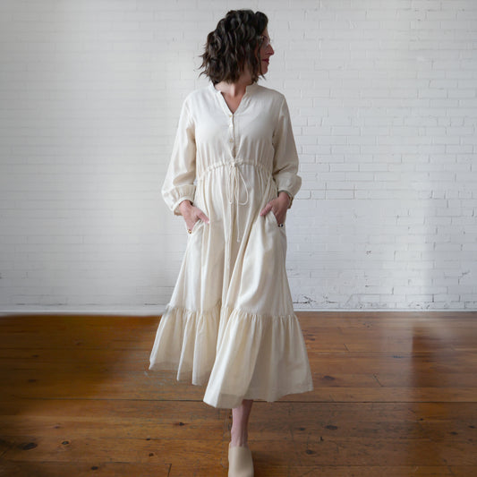 Woman wearing a white dress standing in a room with wooden floors and a white brick wall.