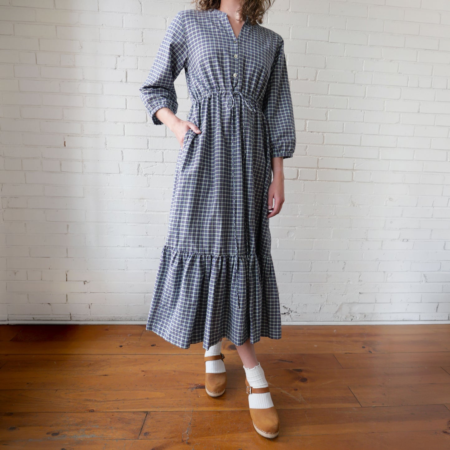 Person wearing a checkered dress standing against a white brick wall.