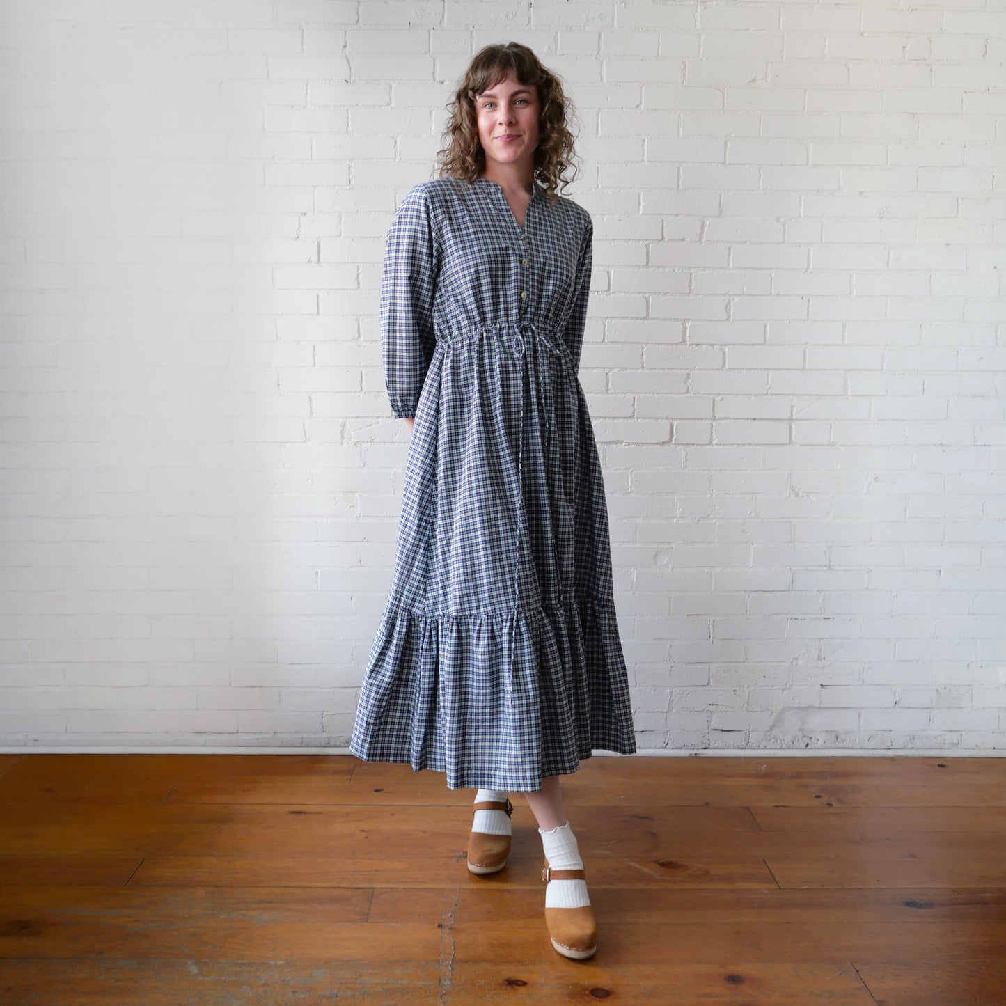 Woman wearing a blue checkered dress standing against a white brick wall.