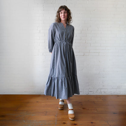 Woman wearing a blue checkered dress standing against a white brick wall.