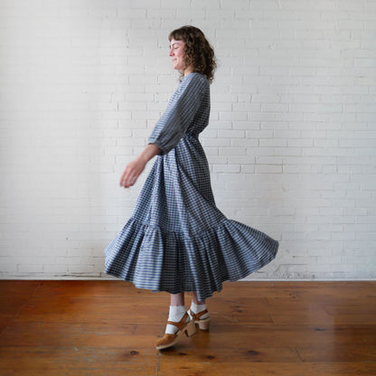 Woman wearing a blue and white checkered dress standing in front of a white brick wall.