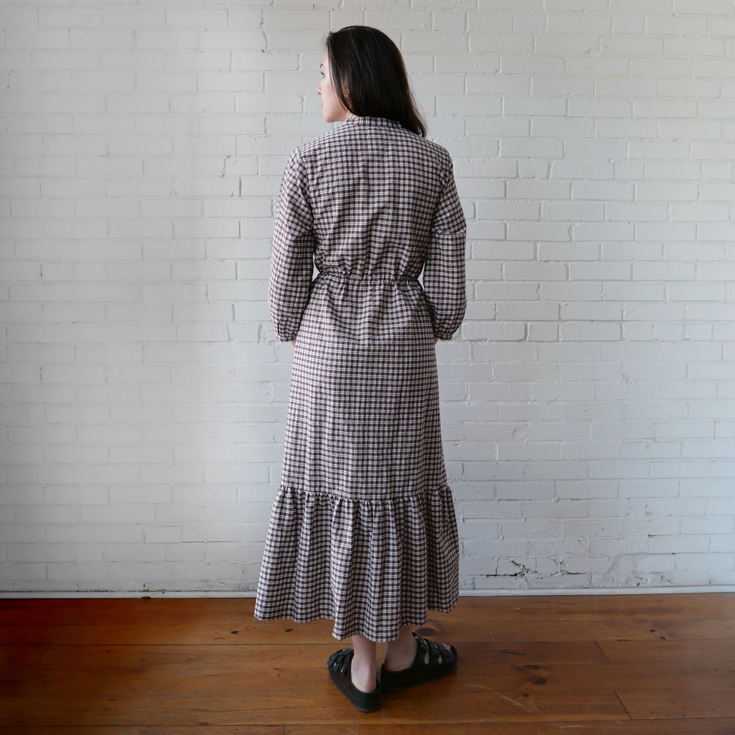 Woman wearing a checkered dress standing against a white brick wall.