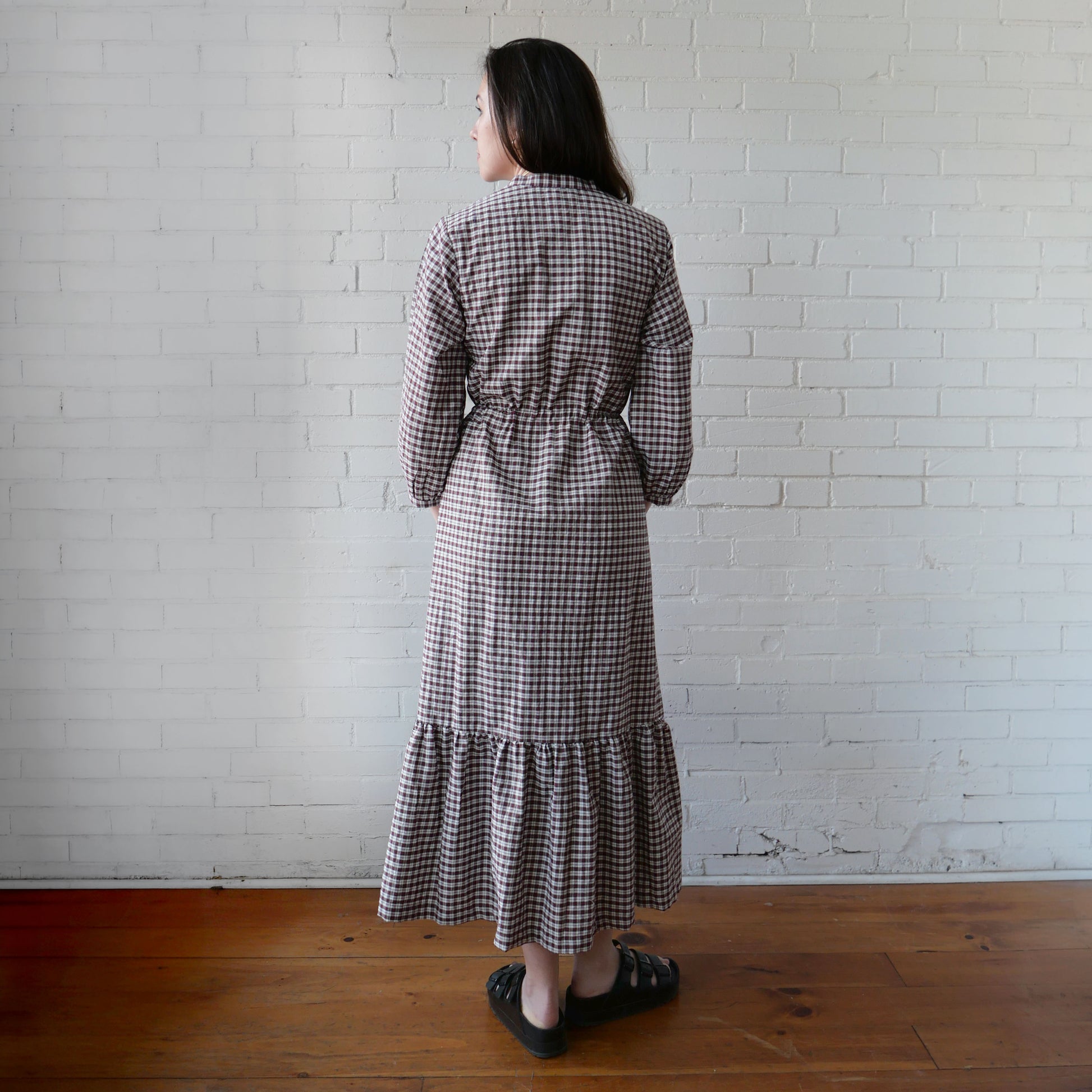 Woman wearing a checkered dress standing against a white brick wall.