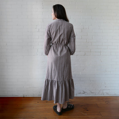 Woman wearing a checkered dress standing against a white brick wall.