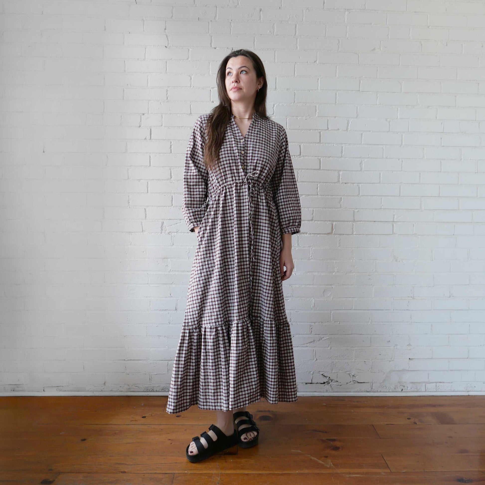 Woman wearing a checkered dress standing against a white brick wall.