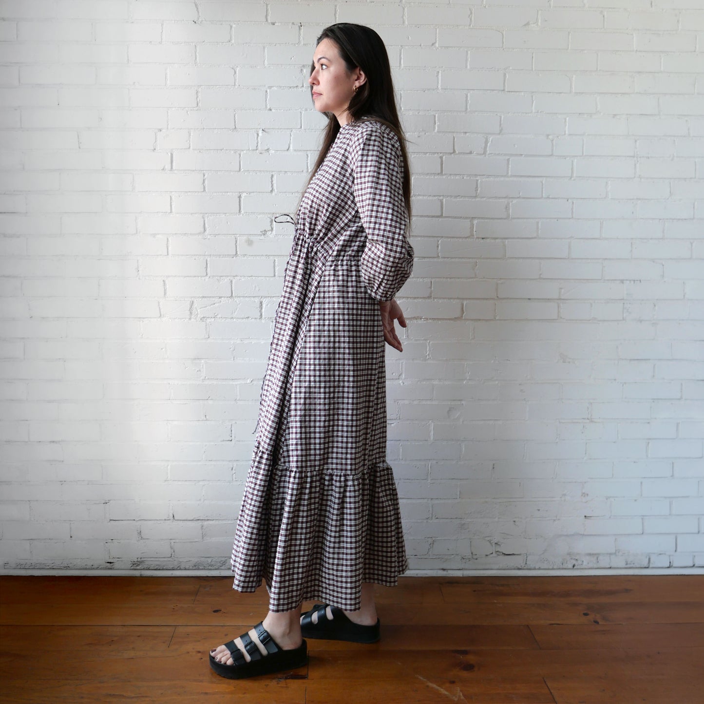 Woman wearing a checkered dress standing against a white brick wall.