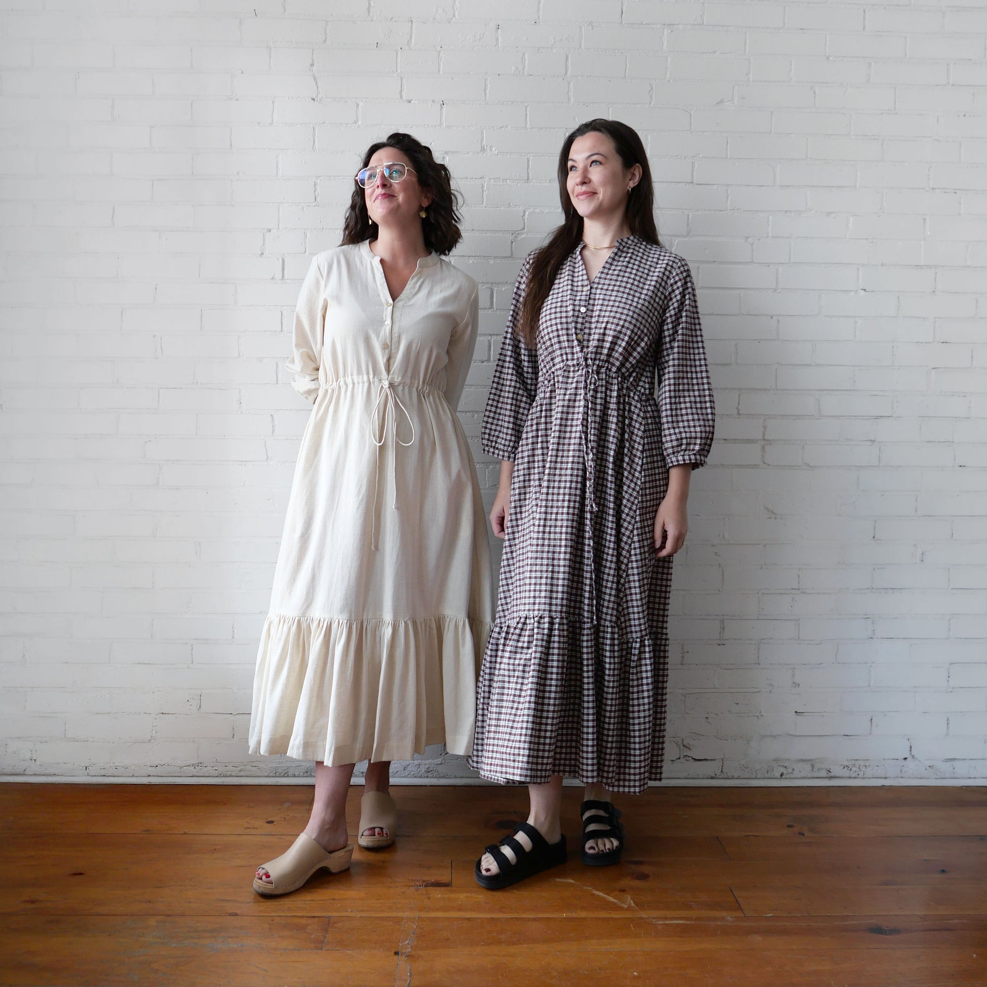 Two women standing side by side against a white brick wall, wearing long dresses.