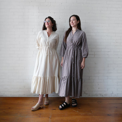 Two women standing side by side against a white brick wall, wearing long dresses.