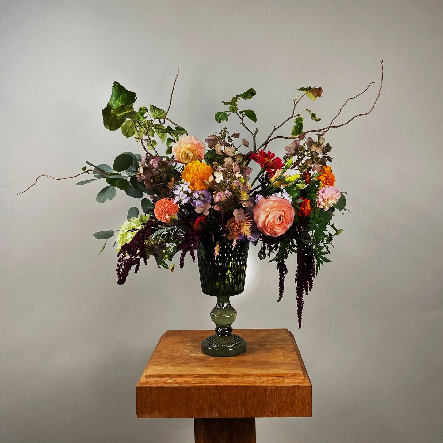 A large bouquet of fresh flowers arranged in a glass vase, placed on a wooden pedestal against a neutral background.
