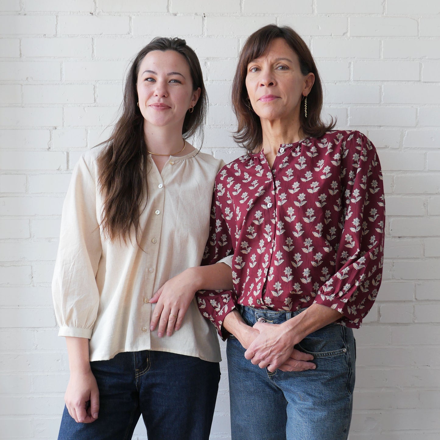 Two women standing side by side against a white brick wall.