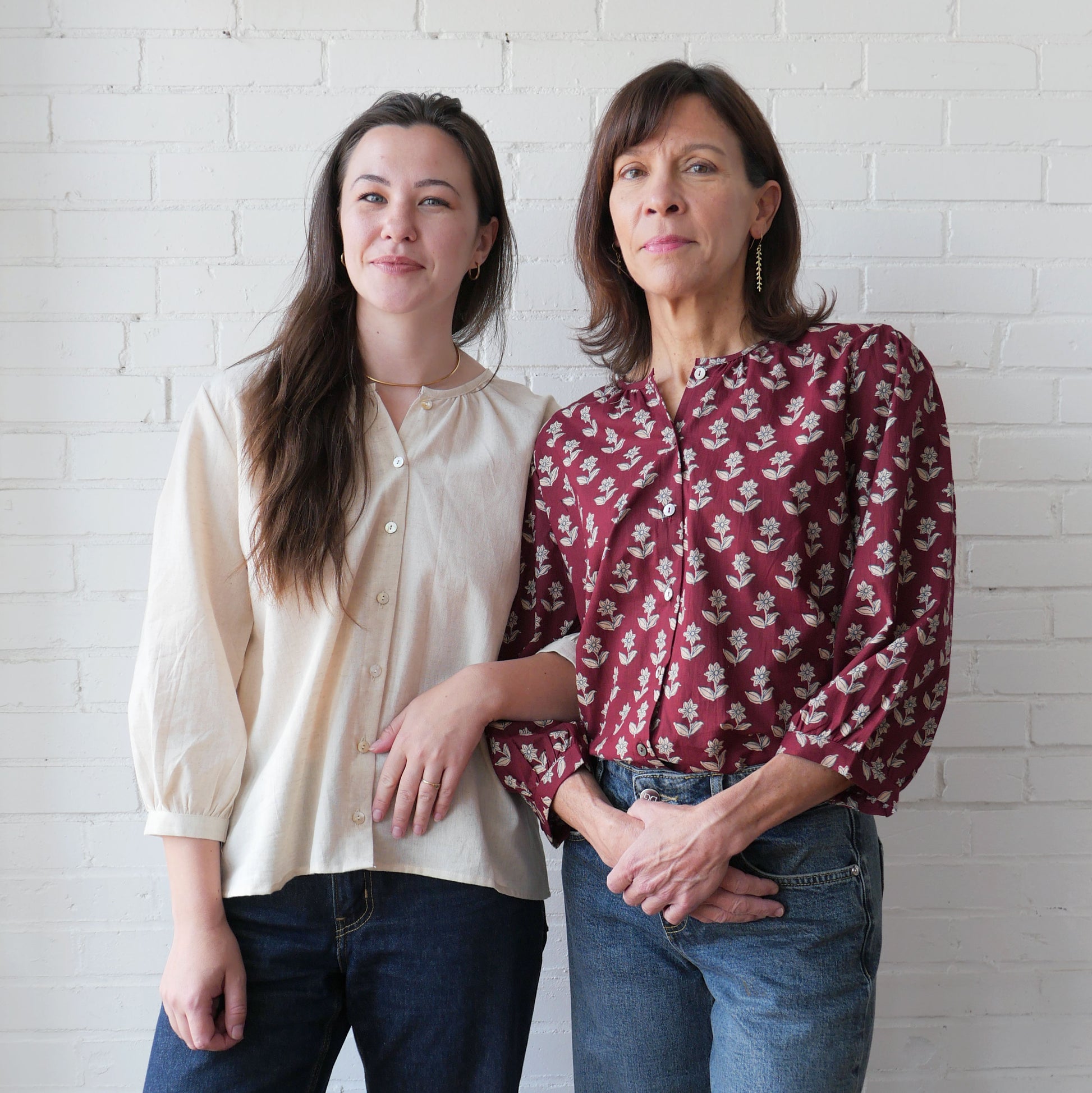 Two women standing side by side against a white brick wall.
