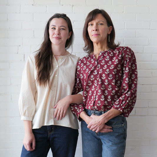 Two women standing side by side against a white brick wall.