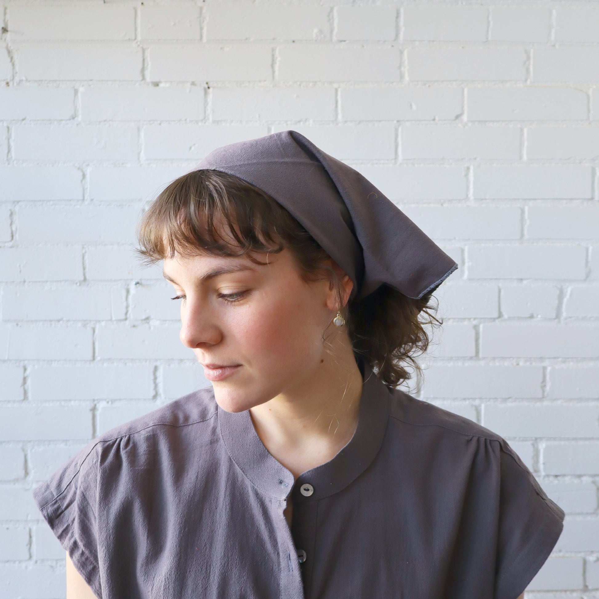 Woman wearing a gray headscarf and shirt against a white brick wall
