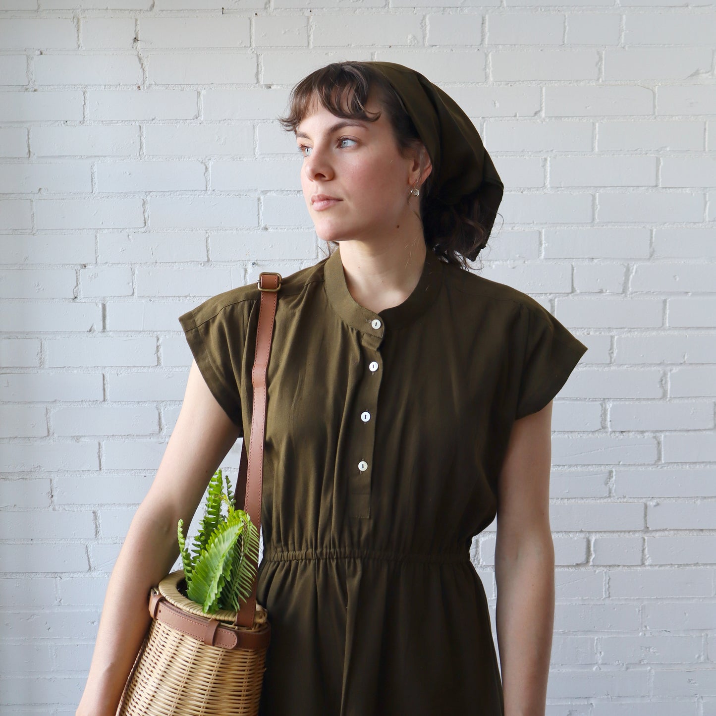 Woman in a green dress holding a woven basket with a plant against a white brick wall.