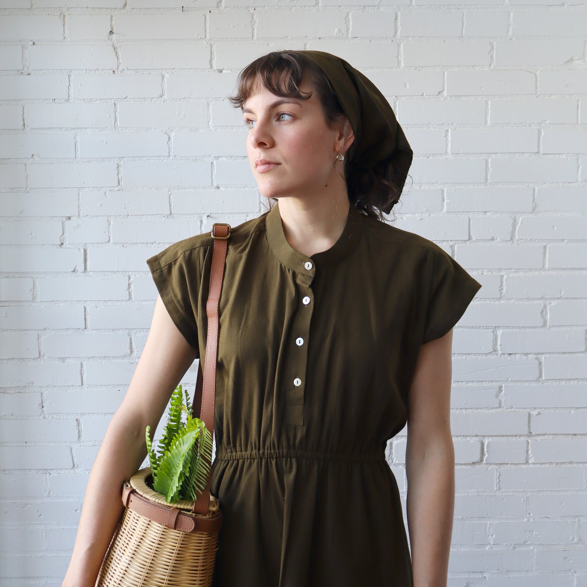 Woman in a green dress holding a woven basket with a plant against a white brick wall.