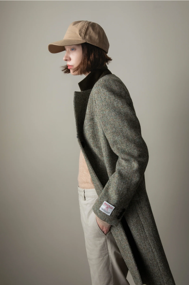 Person wearing a long dark coat, beige cap, and light pants against a plain background