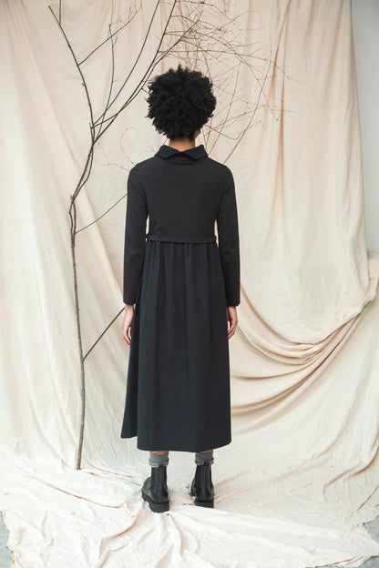 Person wearing a black dress standing against a beige curtain with branches.