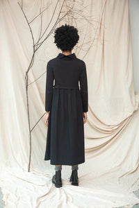 Person wearing a black dress standing against a beige curtain with branches.