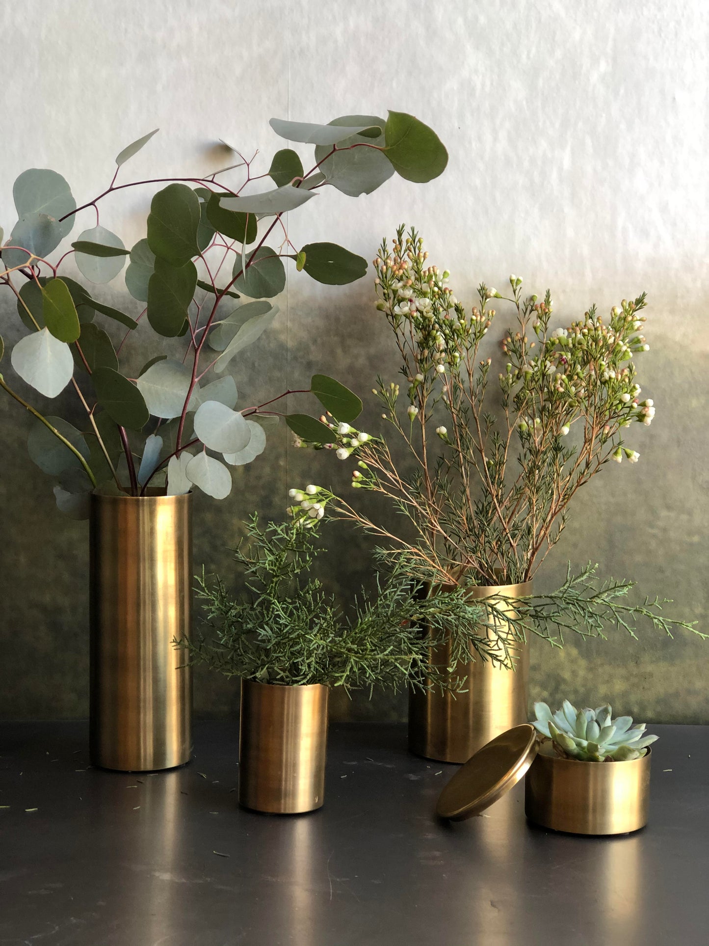 Gold vases with greenery on a dark surface against a textured wall.