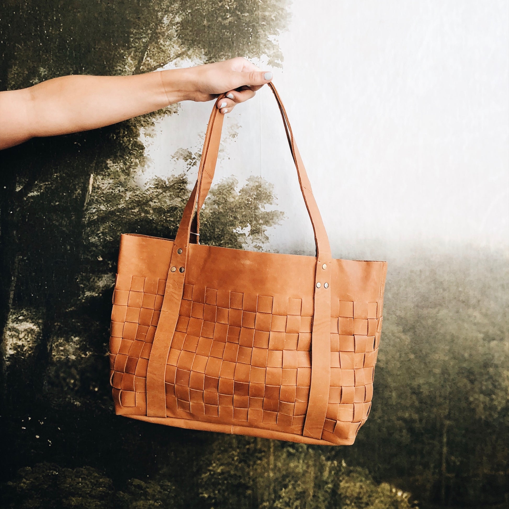 Brown woven handbag held by a person against a textured wall.