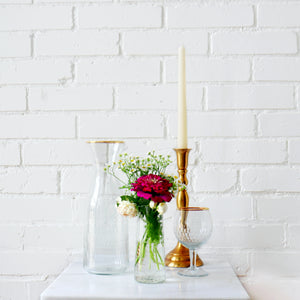 Decorative items including a clear vase with flowers, a gold candlestick, and a glass bowl on a white surface with a white brick wall background.